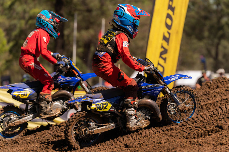 Two young child motocross riders in red racing gear and blue helmets competing on Yamaha YZ65 motorcycles during a kids race on a dirt track.