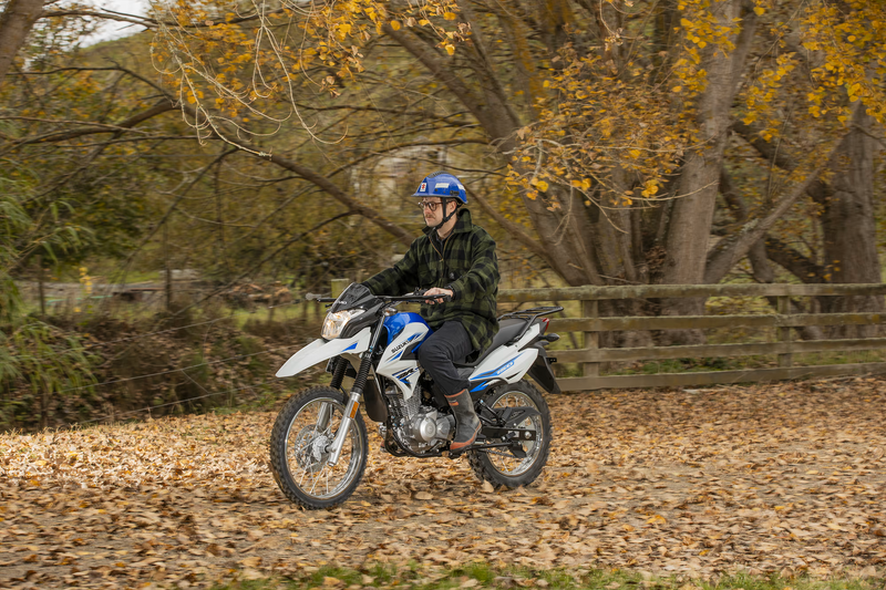 Load image into Gallery viewer, A person wearing a blue helmet and dark plaid jacket rides a white and blue off-road 2026 Suzuki DR 150 motorcycle on a leaf-covered path in an autumn park with trees and a wooden fence.
