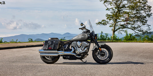 A 2026 Indian Super Chief Dark Horse motorcycle with black saddlebags and chrome exhausts parked on an asphalt road with mountains and trees in the background.