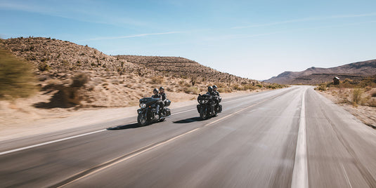 Two 2026 Indian Pursuit Dark Horse motorcycles travel side by side on a paved desert highway with rocky hills and a clear blue sky.