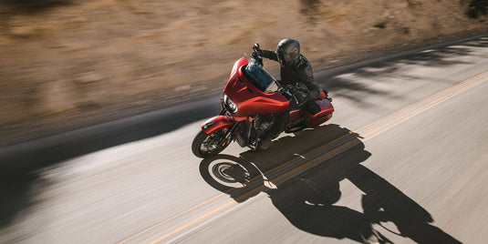 A red 2026 Indian Chieftain Dark Horse motorcycle with a rider in black gear traveling on a paved road with dry brush in the background, captured from a dynamic side angle with motion blur.