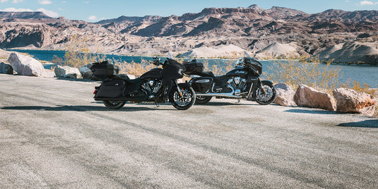 Two black 2026 Indian Pursuit Dark Horse motorcycles parked on a paved road beside a lake, with rocky mountains in the background under a blue sky.
