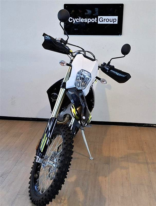 A Triumph TF 450-E dirt bike with a white front panel and black bodywork, displayed on a kickstand in a showroom with a wood floor and a 'Cyclespot Group' sign on the wall.