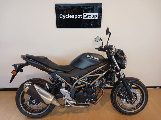 A black 2026 Suzuki SV 650 ABS motorcycle with a treble-cradle frame and chrome exhaust, parked indoors against a beige wall with a 'Cyclespot Group' sign above it.