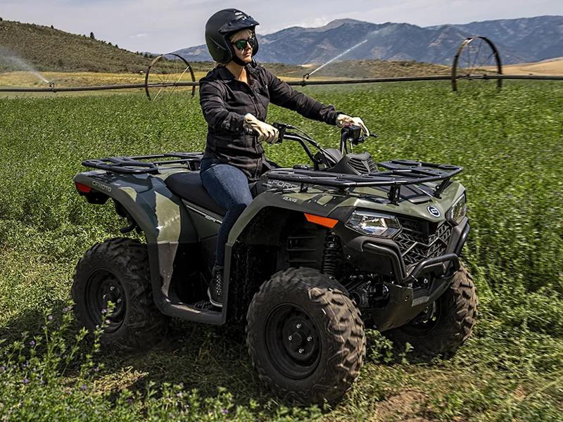 Load image into Gallery viewer, A woman wearing a black helmet and riding gear sits on a 2025 CFMoto CFORCE 400 Hunter Green all-terrain vehicle (ATV) in a green field with mountains in the background.
