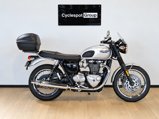 A silver and black Triumph Bonneville T100 motorcycle with a black top case, displayed in profile on a light wood floor against a white wall with a 'Cyclespot Group' sign above it.