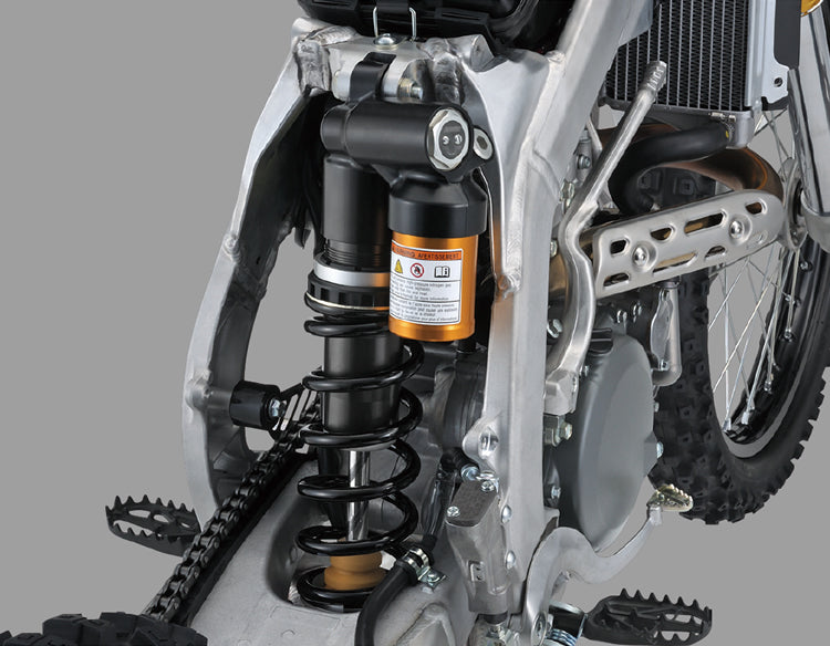 Load image into Gallery viewer, Close-up of a motorcycle's rear suspension system, featuring a black coil spring, black shock absorber, and an orange filter canister, set against a silver frame and tire.
