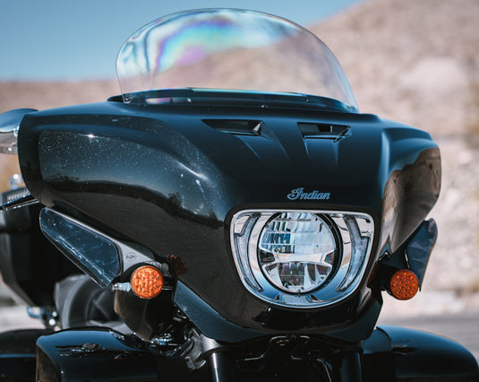 Close-up of the front of a black 2026 Indian Roadmaster Dark Horse motorcycle with a clear windshield, chrome headlight, and amber side markers, photographed outdoors against a blurred desert background.